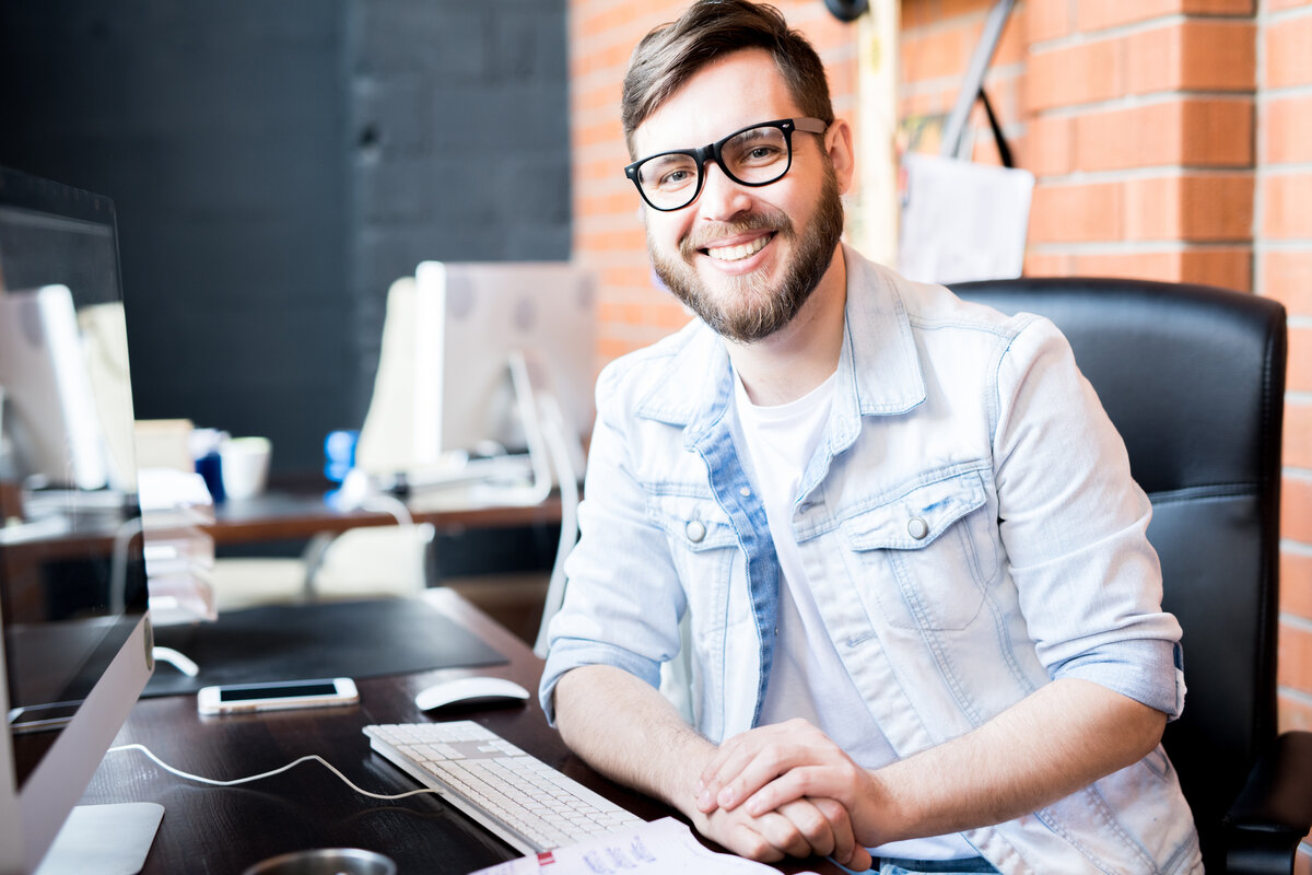 A smiling man sitting at a desk in front of a computer.