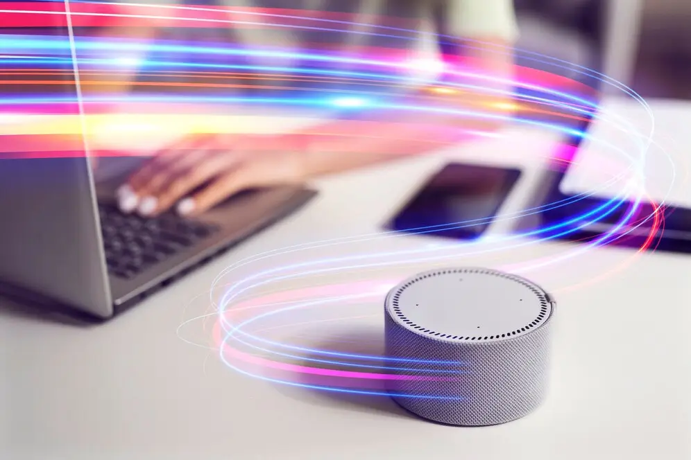 A person typing on a laptop, with a smartphone nearby and a smart speaker in the foreground surrounded by colorful light streaks.